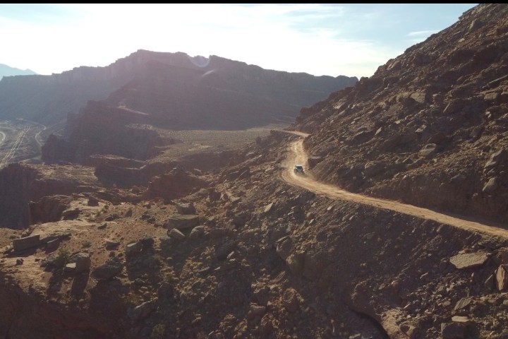 Car on a dirt road winding through rocky desert hills with distant mountains in the background.