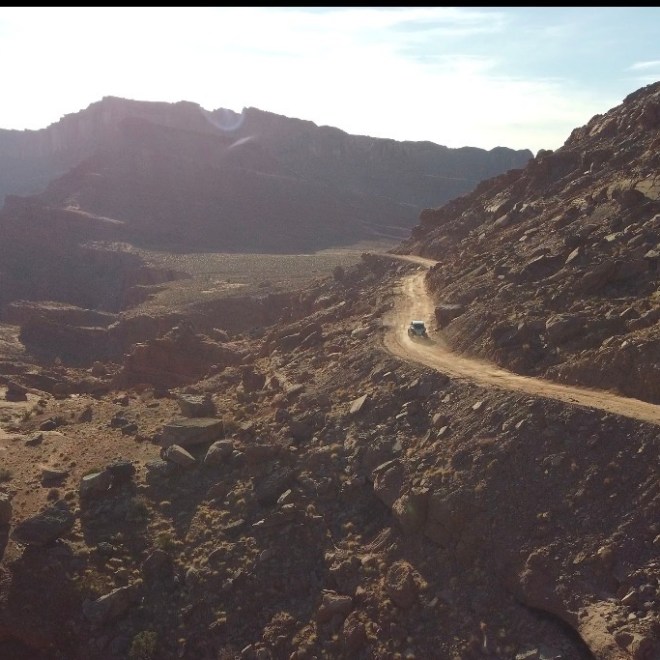 Car on a dirt road winding through rocky desert hills with distant mountains in the background.